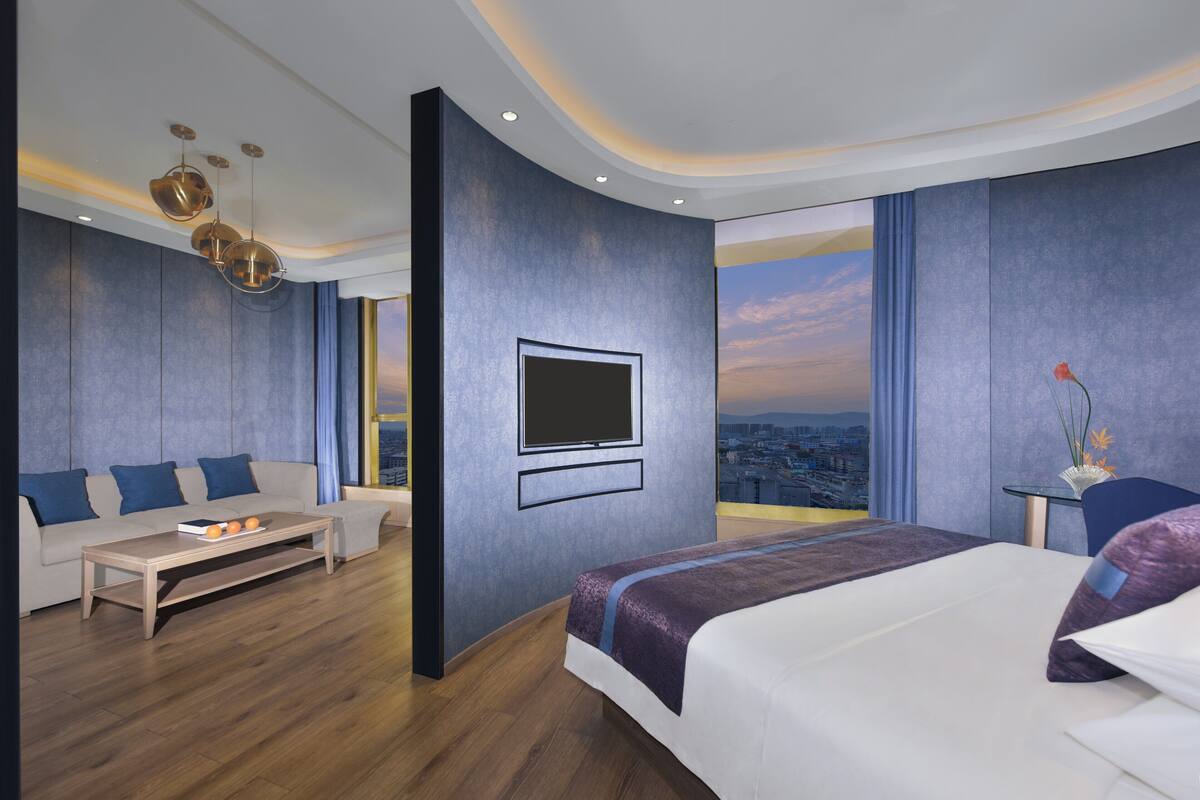 A modern hotel room with a large bed, a sofa, a coffee table, a TV mounted on the wall, and a view of the cityscape.