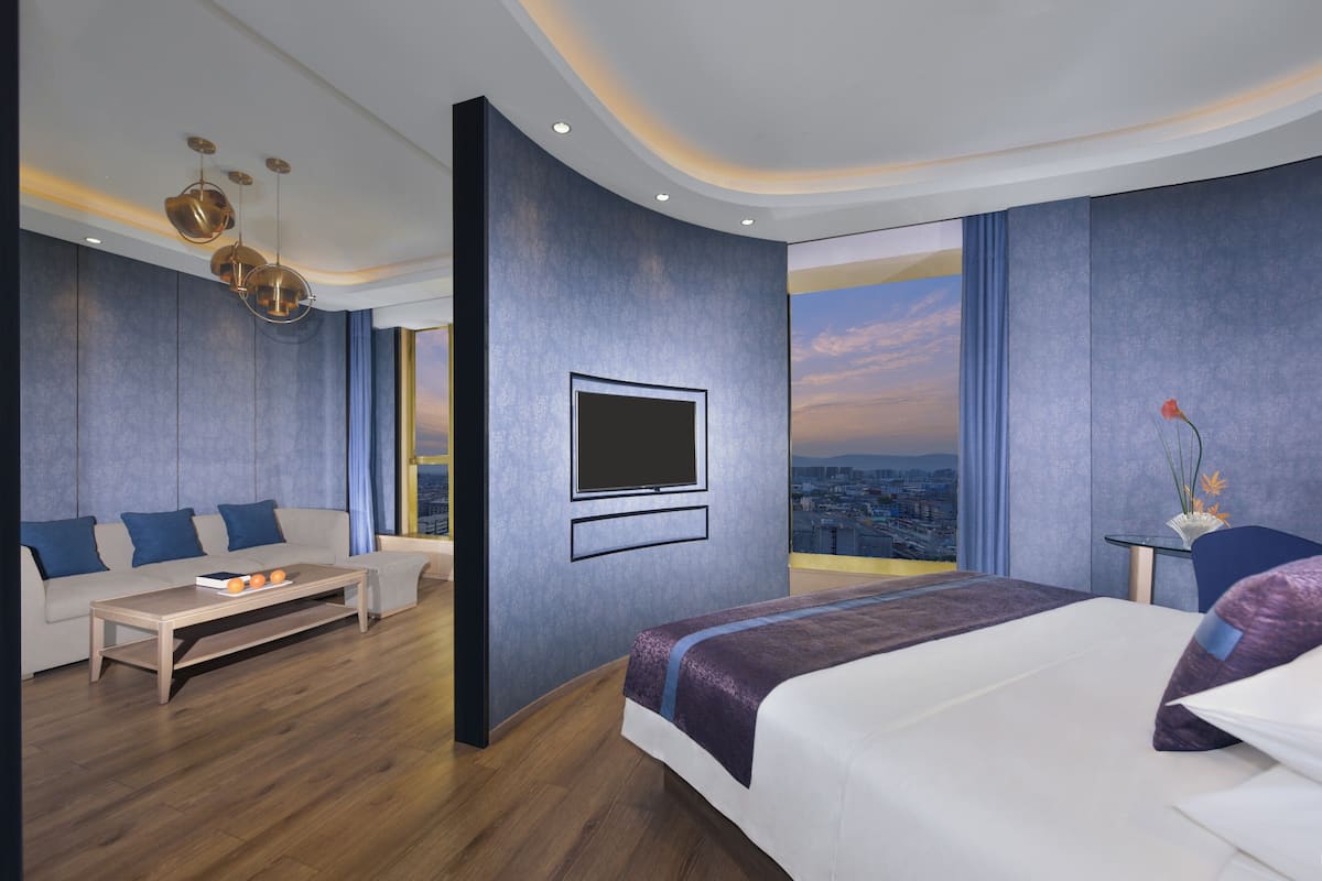 A modern hotel room with a large bed, a sofa, a coffee table, a TV mounted on the wall, and a view of the cityscape.