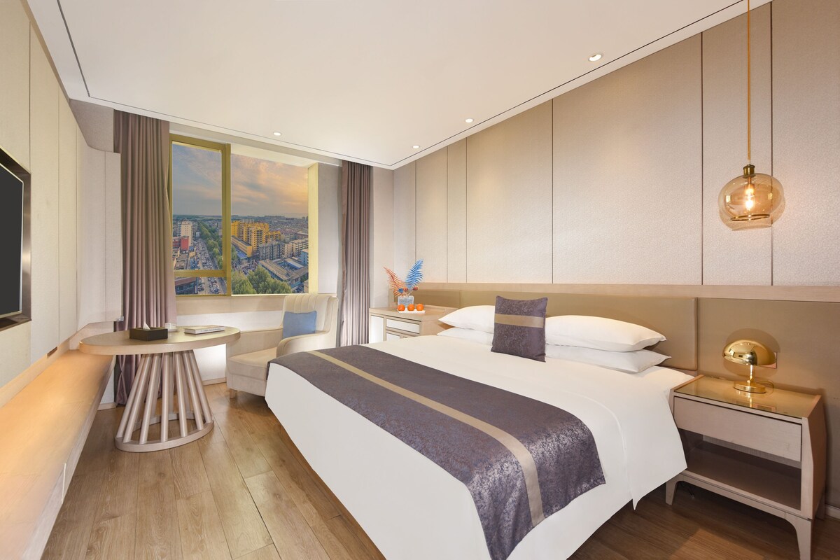 A modern hotel room with a large bed, a bedside table, a nightstand, a desk, a chair, a window with a city view, and a ceiling light fixture.