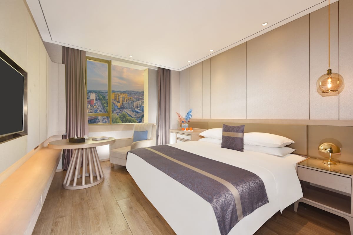 A modern hotel room with a large bed, a bedside table, a nightstand, a desk, a chair, a window with a city view, and a ceiling light fixture.