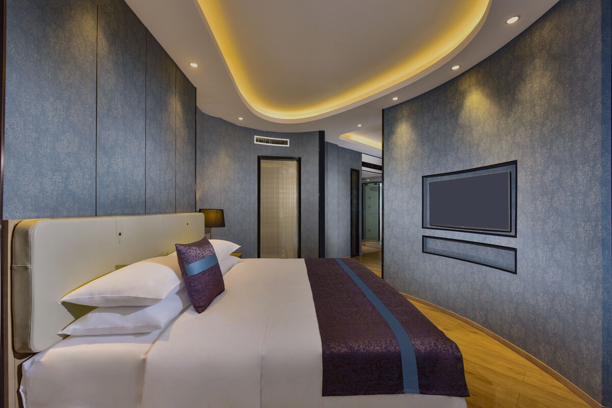A modern hotel room with a large bed, a flat-screen TV mounted on the wall, and a curved ceiling design.