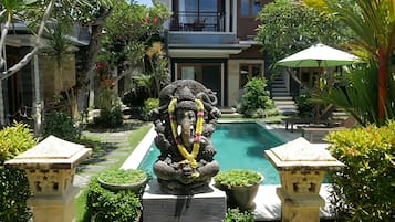 A statue of a deity in a garden with a pool, surrounded by lush greenery and a building in the background.
