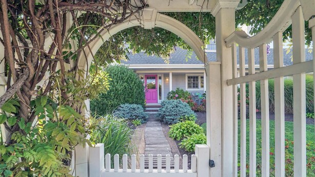 A white wooden gate leading to a house with a pink door, surrounded by lush greenery and a wooden pathway.
