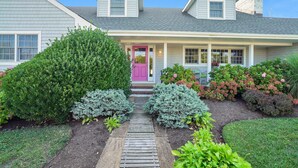 A two-story house with a front porch, a pink door, and a well-maintained garden with various shrubs and a wooden pathway.