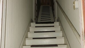 Un escalier étroit, peint en blanc, avec une rampe métallique, menant à un intérieur sombre.