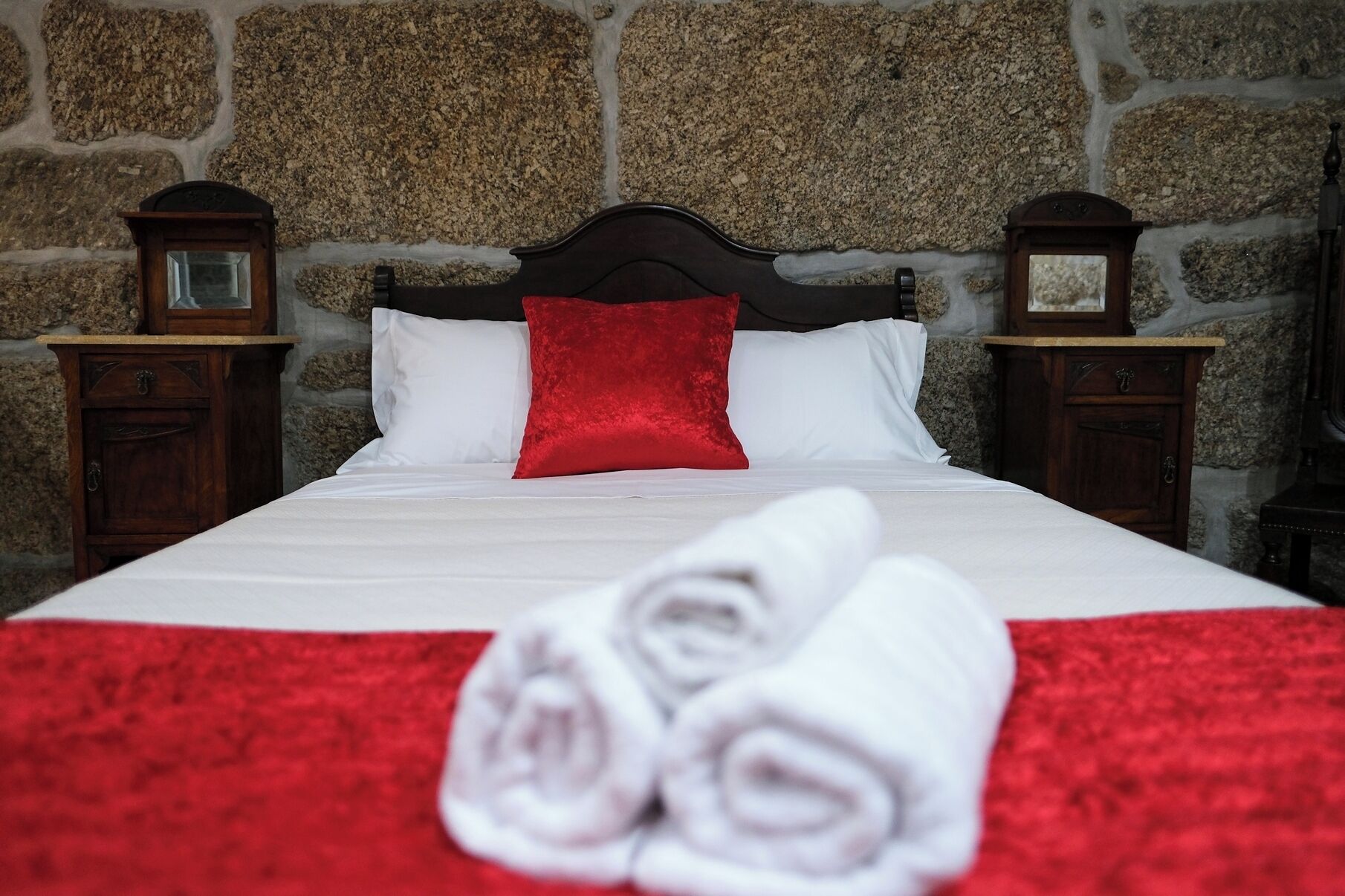 A neatly made bed with white linens and a red pillow, set against a stone wall background.