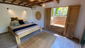 Un dormitorio con una cama de madera, un tapete tejido, un aire acondicionado de pared y una gran puerta de madera que da a un espacio exterior.