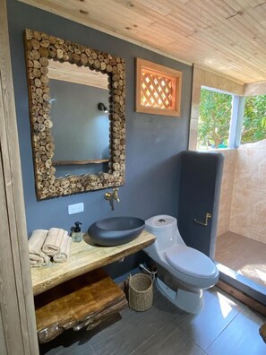 A bathroom with a wooden-framed mirror, a wooden vanity with a basin, a toilet, and a window with a view of greenery. - cabañas bari  (Moñitos)