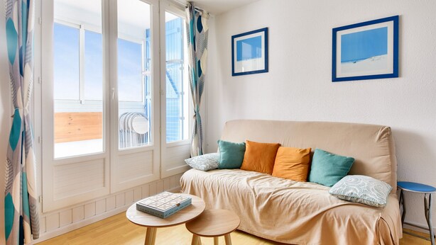 A room with a beige sofa, two framed pictures, a small round table, and a view of the sea through large windows. - 250m from the beach - Loggia - Flat for 4 (Saint-Hilaire-de-Riez)