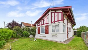 Une maison à deux étages avec un toit rouge et des murs blancs, un petit jardin avec une pelouse et une table entourée de chaises, et une clôture en bois.