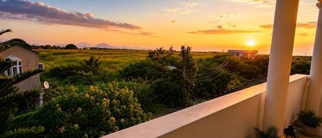 A balcony with a view of a sunset over a field, a house, and some vegetation.