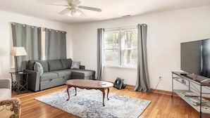 Ein Wohnzimmer mit einem Ecksofa, einem Couchtisch, einem Fernseher auf einem Lowboard und einem Fenster mit Vorhängen.