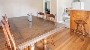 Ein Essbereich mit Holztisch und -stühlen, eine Küchenzeile mit Kühlschrank und ein Tresenstuhl, sowie ein Gang, der zu einem weiteren Raum führt.