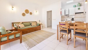 Un salon moderne comprenant un canapé, des chaises en bois, un tapis et un coin cuisine avec des placards et des appareils électroménagers.