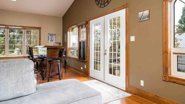 A spacious living room with a gray sofa, wooden flooring, and large windows offering a view of the outdoors. - JJ’s small town country retreat     (Badjeros)