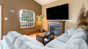 A living room with a fireplace, a large window, and a flat-screen TV mounted on the wall. - JJ’s small town country retreat     (Badjeros)