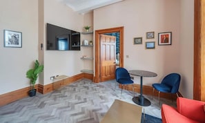 Une chambre d’hôtel avec un parquet en chevrons, une porte en bois, une table ronde avec deux chaises, un canapé rouge et une plante en pot.