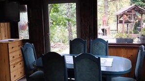 A dining area with a wooden table and chairs, a sofa with a green pillow, and a view of the outdoors through large windows. - Cottage in the Picturesque Beskid Maly Mountains (Porabka)