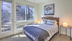 Une chambre dotée d’une grande fenêtre, d’un ventilateur de plafond, d’un lit avec une tête de lit en bois et d’une table de chevet avec une lampe.