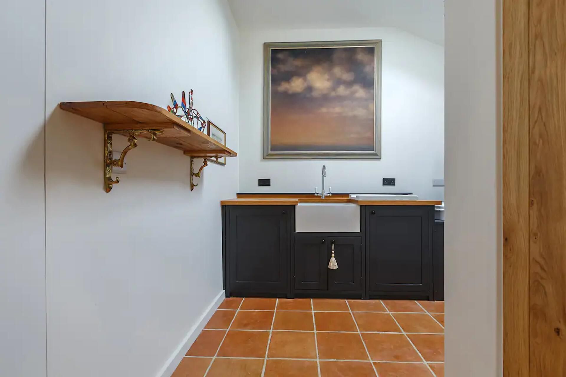 A kitchen with a wooden shelf, a painting of a sunset, and a tiled floor.
