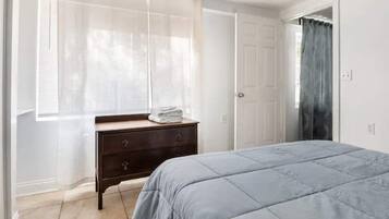 Ein Schlafzimmer mit Bett, Kommode und einem Fenster mit transparenten Vorhängen.