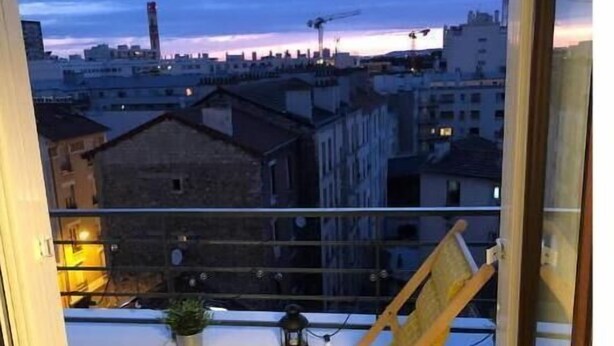 Ein Balkon mit einem Holzstuhl und einem Blumentopf, mit Blick auf eine Stadtlandschaft im Abendlicht.