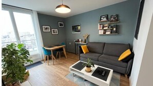 Ein modernes Wohnzimmer mit grauem Sofa, einem Couchtisch und einem Esstisch mit Stühlen. An der Wand hängen gerahmte Bilder und auf dem Boden steht eine Pflanze.