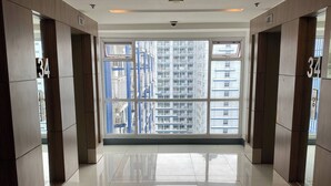 A spacious hallway with reflective tiled floors, large windows offering a view of high-rise buildings, and numbered doors on the walls. - Grass Residence T3 34th Floor (Quezon City)