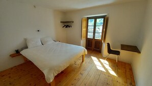 A bedroom with a wooden floor, a bed with white bedding, a chair, a small shelf, and a window with a view of buildings. - Room-"casa Coerente" Cavergno - Rooms #1 (Cevio)