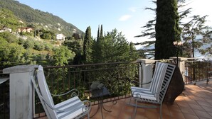 Ein Balkon mit zwei weißen Stühlen und einem Tisch, mit Blick auf eine gebirgige Landschaft mit Häusern und Bäumen.