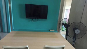 A room with a teal wall, a flat-screen TV mounted above a wooden table, two white chairs, and a standing fan. - Grass Residence Tower ii 2011 (Quezon City)