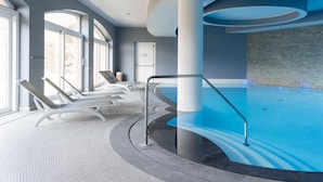 Una piscina interior moderna con diseño curvo, sillones de descanso y vistas al exterior a través de amplios ventanales.