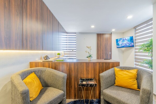 Un área de recepción con un mostrador de madera, dos sillones con cojines amarillos, un televisor de pantalla plana en la pared y una planta en maceta.