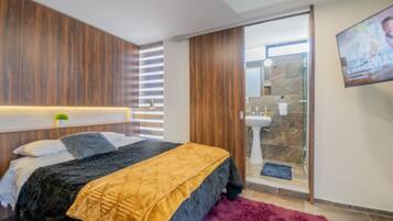 A modern bedroom with a bed, a TV mounted on the wall, and a bathroom with a glass shower enclosure.