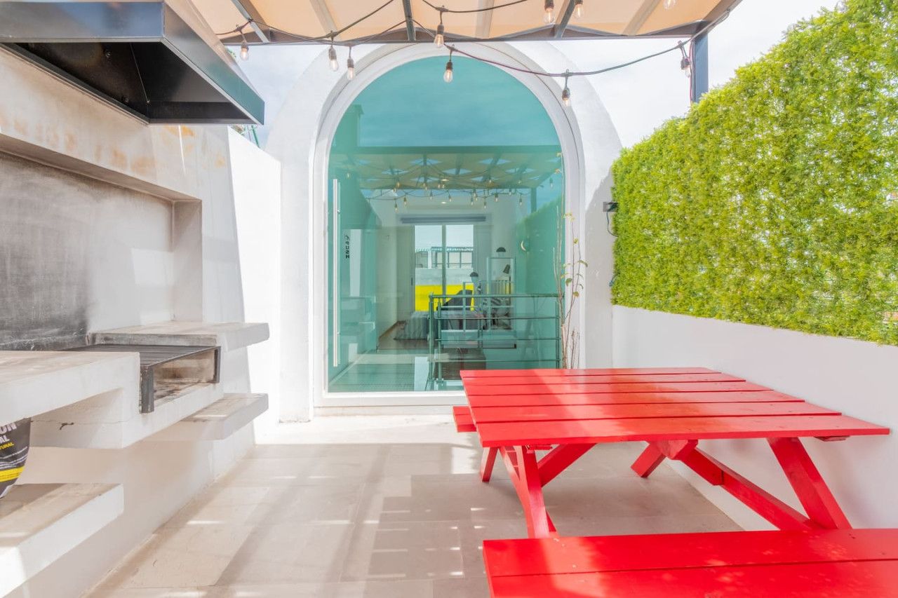 Een terras buiten met een rode tafel en een barbecue, een glazen deur naar een kamer en een groene haag.