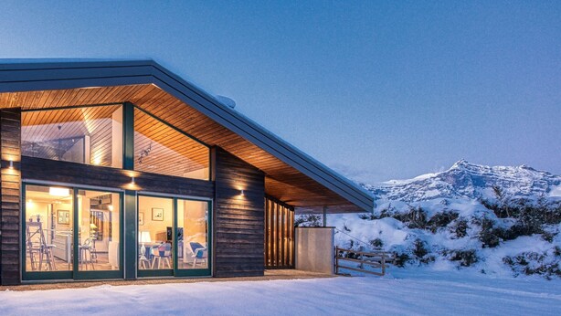 Een modern gebouw met grote glasramen en een houten gevel, op de achtergrond een besneeuwde berg.