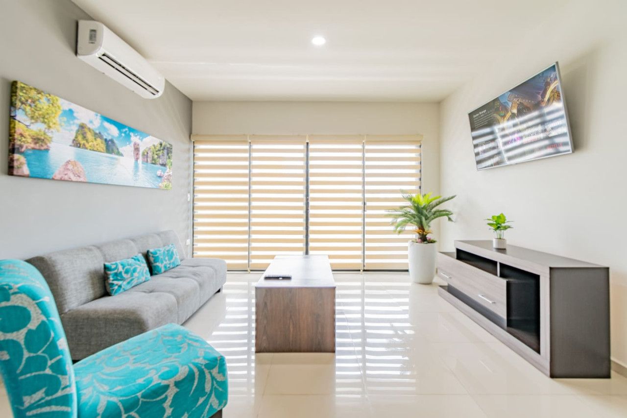 A modern living room with a grey sofa, a wooden coffee table, a TV stand, and a large window with blinds.