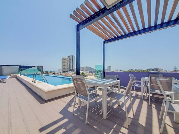A rooftop pool area with a view of the city skyline, outdoor seating, and a pergola structure.