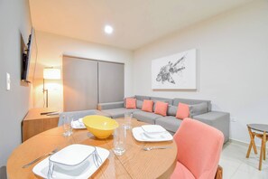 A modern living room with a sofa, a dining table set with plates and glasses, a TV mounted on the wall, and a lamp.