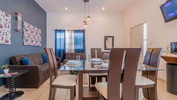 A modern living room with a glass dining table, brown sofas, and a TV mounted on the wall.