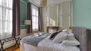 A neatly made bed with a grey and white patterned duvet, a grey headboard, and white pillows. There is a small table with a lamp and a decorative item on it. - La Villa Authentique by Villas Signature (Bourbourg)