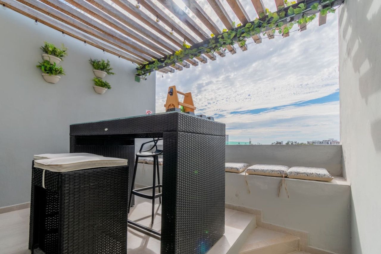 Un balcón con una mesa y sillas de ratán negro, plantas en macetas y vista al mar.