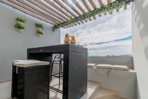 A balcony with a black wicker table and chairs, potted plants, and a view of the sea.
