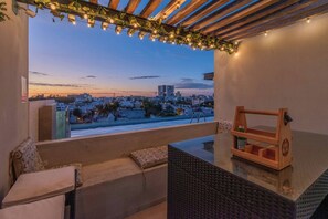 A balcony with a view of a cityscape at dusk, a wooden bench, a wicker table, and a small wooden shelf with a bottle. - Penthouse Con Terraza en Zona Dorada (Mazatlán)