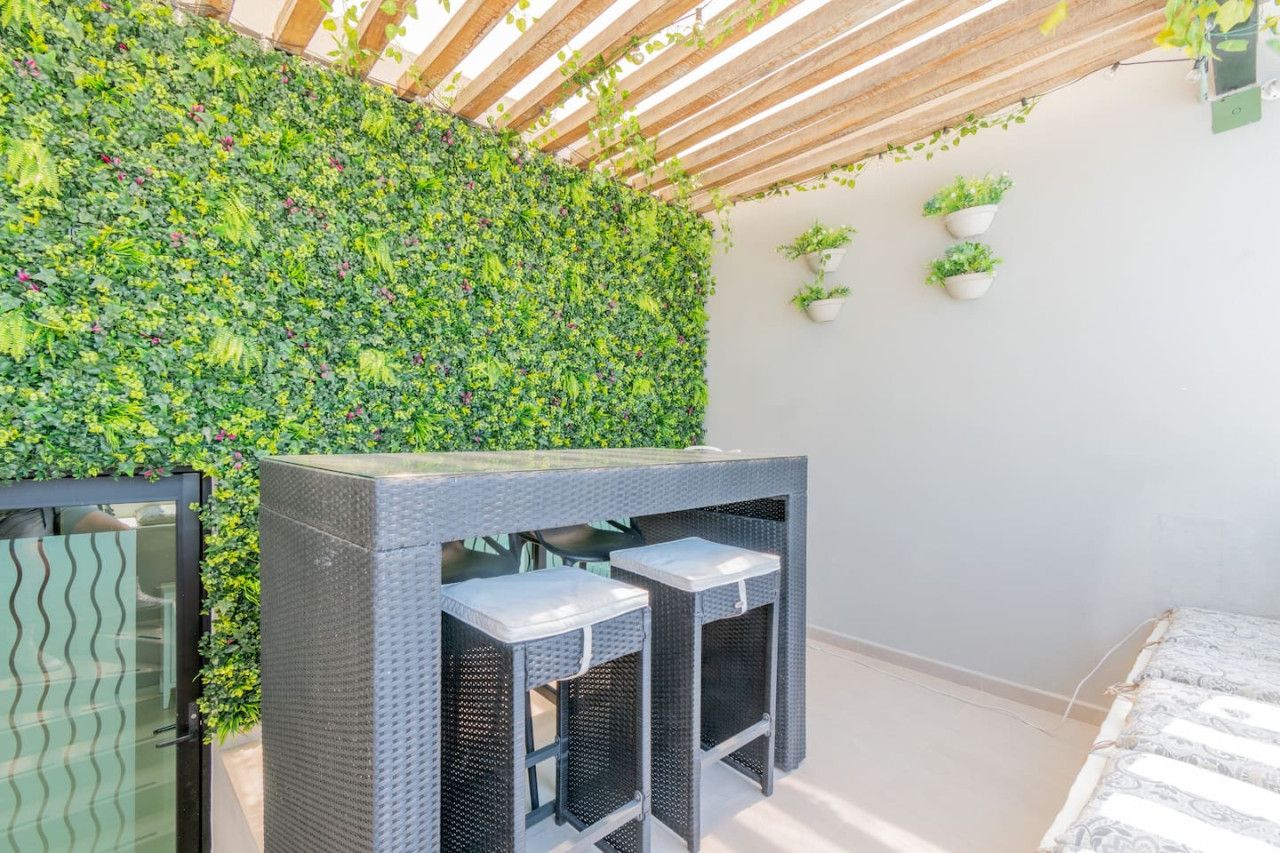 A room with a green wall covered in foliage, a wicker table with two chairs, and a bed with patterned bedding.