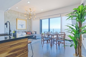A modern kitchen with a central island, dining area with wooden chairs, a large window with a view, and a chandelier. - Depa en Malecon con Vista al Mar (Mazatlán)