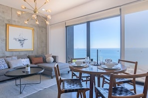 Un salon moderne avec un canapé, une table basse et un coin repas avec des chaises. Une grande fenêtre offre une vue sur la mer.