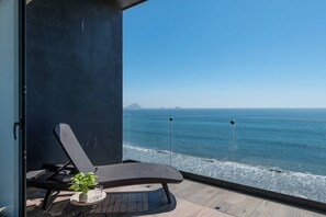 A balcony with a view of the ocean, a lounge chair, a small table with a plant, and a glass door.