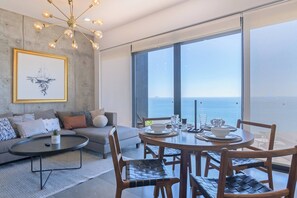 A modern living room with a sofa, coffee table, and dining table set for four, large windows with a view of the sea, and a chandelier.
