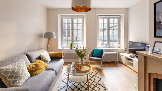 A modern living room with a grey sofa, a coffee table, and a TV. There are two windows with white frames and a view of buildings outside. - Chez Léon - Appt à 500m des plages (Saint-Malo)